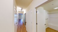 This single-floor unit offers a thoughtfully designed layout with consistent warm hardwood-style flooring that flows seamlessly throughout every room. The spacious living area is the centerpiece of the unit, featuring clean white walls, a ceiling with subtle architectural beam detailing, and a large floor-to-ceiling window that frames expansive city views, filling the space with abundant natural light. Adjacent to the living area, the kitchen is well-appointed with warm yellow cabinetry, light stone countertops, and a full suite of stainless steel appliances including a refrigerator, electric range, built-in microwave, and dishwasher. Track lighting overhead ensures excellent task illumination. A hallway connects the living spaces to the more private areas of the unit, lined with multiple doors providing ample storage and closet space. The bathroom is clean and functional, featuring a dual-sink vanity with light stone countertops, warm wood cabinetry with brushed metal hardware, a large mirror, and a full soaking tub with shower. The unit's neutral color palette of warm creams and whites creates a bright, welcoming ambiance throughout.
