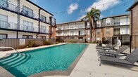 This inviting outdoor pool area is located in Metairie, Louisiana, and serves as a central communal amenity within a multi-story residential complex. The space is defined by its generously sized swimming pool with clear, blue-green water, framed by a wide concrete pool deck that provides ample room for lounging and socializing. A row of modern chaise lounge chairs lines one side of the deck, offering comfortable spaces to relax poolside.

The area is enclosed by a sleek wrought-iron safety fence, providing security while maintaining an open, airy feel. Overhead, strands of globe string lights are strung across the courtyard, adding a warm, festive ambiance ideal for evening gatherings. Lush tropical landscaping, including a tall palm tree and potted greenery, adds a resort-like character to the space.

The pool is flanked on multiple sides by the surrounding multi-story buildings, which feature classic exposed brick facades, arched windows, white-trimmed balconies with wrought-iron railings, and decorative gas-style wall lanterns — all lending a distinctly New Orleans-influenced architectural charm. Additional seating with tables is visible on a covered terrace nearby. The courtyard layout creates a sense of enclosure and privacy, making this pool area a welcoming outdoor retreat.