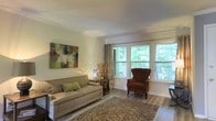 This single-floor space offers a welcoming and well-appointed living area designed with comfort and style in mind. The sitting room is a standout feature, characterized by a large bank of windows that flood the space with abundant natural light and frame lush, green tree views beyond, creating a seamless connection to the surrounding outdoor environment. White walls and crisp trim work throughout contribute to a clean, airy aesthetic.

The living area is finished with wood-style flooring that adds warmth and texture underfoot, complemented by a woven area rug that grounds the seating arrangement. Furnishings include a tufted neutral-toned sofa accented with green throw pillows and a rich burgundy wingback chair, lending a classic yet approachable character to the room. A decorative abstract canvas painting adds a pop of color to the wall, while a sculptural table lamp and dried floral arrangement introduce organic, layered details.

Curtain panels frame the windows elegantly without obstructing the natural light. The overall ambiance of the space is calm, bright, and inviting, with thoughtful styling that balances functionality and visual appeal throughout.
