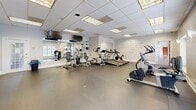 Located at 3800 Michael Blvd in Mobile, Alabama, this single-floor fitness center offers a thoughtfully designed exercise space for residents or members seeking a convenient and well-appointed workout facility. The exercise room is generously sized, featuring durable dark flooring suited for high-traffic use, a drop-panel ceiling with recessed fluorescent lighting, and neutral gray walls that create a clean, motivating atmosphere. The space is equipped with a variety of cardio machines, including treadmills, ellipticals, and a stationary bike, alongside a multi-station strength training unit and a free-weight rack. Wall-mounted televisions are positioned for easy viewing during workouts. Natural light filters in through shuttered windows, enhancing the open feel of the room. Inspirational quotes from notable figures are painted directly on the walls, adding an uplifting and encouraging ambiance. A large mirror along one wall visually expands the space and supports exercise form. Adjacent to the main workout area, a pass-through kitchenette is outfitted with dark wood cabinetry, a stainless steel refrigerator, and a countertop bar, providing a functional space for refreshments. A glass-paneled wood door offers a welcoming entry point to the facility.