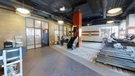 This fitness center combines industrial design with functional workout space. The exposed ceiling reveals ductwork and piping finished in dark tones, contributing to an urban, loft-inspired aesthetic. Pendant lighting and recessed fixtures provide adequate illumination throughout the space. A distinctive red brick column serves as a striking architectural focal point, complemented by horizontal brick-patterned wall accents that add visual interest. The flooring transitions between smooth concrete and patterned carpet tiles, defining different activity zones within the space. Large glass-paneled partition walls and a glass entry door allow natural light to flow in while maintaining a sense of openness and visibility. The cardio zone is well-equipped with stair climbers, elliptical machines, and additional cardio equipment arranged on the carpeted section. An open area with smooth flooring provides space for stretching or floor exercises. The overall ambiance is clean, spacious, and functional, blending modern gym amenities with an architecturally distinctive environment suited for a variety of fitness activities.