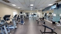 This fitness center offers a thorough and well-organized workout environment designed to support a wide range of exercise routines. The space is laid out across a single floor and divided into distinct functional zones, each dedicated to a specific type of training.

The strength training area is generously stocked with a variety of cable machines, multi-functional weight stations, free weights, barbells, and adjustable benches. A full wall of mirrors lines one side of the room, allowing users to monitor their form while training. The free weight section includes a complete dumbbell rack alongside flat and incline benches, providing ample options for resistance workouts.

A separate cardio zone houses a row of modern treadmills and elliptical machines positioned near natural light from windows along the walls, creating a bright and energizing atmosphere. Overhead, a dropped ceiling with recessed panel lighting ensures consistent illumination throughout the facility.

The flooring consists of durable rubber matting, offering both safety and comfort underfoot. Wall-mounted televisions are positioned throughout the space for entertainment during workouts. The neutral-toned walls and open layout contribute to a clean, functional ambiance. Overall, this fitness center is thoughtfully arranged to accommodate users of all fitness levels with ease and efficiency.