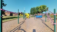 Located at 4187 Beechwold Dr in Columbus, Ohio, this outdoor recreational space offers a vibrant and well-equipped playground set within a generous open-air environment. The grounds are surfaced with wood chip safety material, providing a cushioned play area beneath the various structures. A standout feature is the bright yellow spiral slide, which serves as a central focal point of the playground. Surrounding it are multiple play structures finished in bold green, purple, and blue tones, including overhead horizontal bars designed for climbing and upper-body exercise, as well as a blue tunnel crawl structure that encourages imaginative and active play. The open layout of the space allows for easy visibility across the entire play area. Mature trees line the perimeter, offering partial shade and a natural backdrop that softens the setting. The surrounding lawn is well-maintained, providing additional open space for free play and recreation. A streetlight is present on-site, suggesting the space may be accessible during early evening hours. Nearby brick buildings are visible in the background, situating the playground within an established neighborhood context. Overall, this space presents a functional, colorful, and accessible outdoor play environment suited for children of various ages.