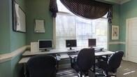 This single-floor space functions as a dedicated computer workroom, designed to accommodate multiple users simultaneously. The room is finished with deep green walls that lend a professional yet comfortable atmosphere, complemented by framed informational signage, including an Integral-branded display, mounted prominently on the wall.

Three computer workstations are arranged along a continuous desk surface that runs the length of the room, each equipped with a monitor, keyboard, and mouse. The setup supports side-by-side use, making it well-suited for collaborative work or individual tasks in a shared environment. Ergonomic task chairs positioned at each station provide functional seating for extended use.

A large bay window dominates one wall, fitted with horizontal blinds that allow users to control natural light levels throughout the day. Decorative black and gold polka-dot valances are draped above the window, adding a touch of style to the otherwise utilitarian space. The window allows considerable daylight to fill the room, reducing reliance on artificial lighting.

The flooring is covered in low-pile carpet, contributing to a quieter working environment. Overall, the space is organized, functional, and well-lit, making it a practical setting for computer-based work.