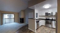 This single-floor home offers an open and functional layout that seamlessly connects the kitchen and living areas. The kitchen is outfitted with white cabinetry, dark countertops, and stainless steel appliances — including a dishwasher, range, and built-in microwave — all set atop wood-look flooring that adds warmth and durability. A peninsula with a sink provides a natural division between the kitchen and the rest of the living space, enhancing workflow and accessibility. Natural light filters in through a window above the sink, keeping the kitchen bright and inviting. The living area features plush carpeting underfoot and is anchored by a tile-framed fireplace with a bold black insert, which serves as a striking architectural centerpiece. Vaulted ceilings rise dramatically over the living room, creating a sense of openness and volume that contrasts with the more intimate kitchen area. Neutral tones throughout — soft beiges and grays — provide a versatile canvas that suits a variety of décor styles. A sliding glass door in the living area hints at outdoor access, further enhancing the sense of light and connection to the exterior. Overall, the space blends practical design with comfortable living.