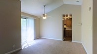 This single-floor residence offers a well-designed layout that balances openness with functionality. The living area serves as the heart of the home, immediately drawing attention with its vaulted ceiling that adds a sense of height and airiness to the space. A ceiling fan is centrally mounted, providing both comfort and a subtle design accent. Plush wall-to-wall carpeting in a neutral tone runs throughout, contributing to a warm and inviting atmosphere. Large sliding glass doors fitted with vertical blinds line one wall, allowing generous natural light to flood the interior and offering access to the outdoors. The open passageway connecting the living area to the kitchen creates a seamless flow between the two spaces. The kitchen features warm, dark wood cabinetry with ample storage, track lighting overhead, and dark appliances that provide a clean contrast to the lighter surroundings. The neutral color palette throughout — from the beige walls to the light carpet — creates a cohesive, understated aesthetic that is both versatile and easy to personalize. The textured ceiling adds a subtle finishing touch to the overall design of this practical and comfortable single-floor home.