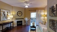 This single-floor residence offers a warm and inviting atmosphere highlighted by a well-appointed living area. The living room serves as the centerpiece of the space, featuring rich, dark hardwood flooring that anchors the room with a sense of elegance and warmth. A tile-surround fireplace with a sleek dark mantle provides a striking focal point, ideal for relaxing evenings.

Warm golden-toned walls complement the natural light that filters in through a glass patio door dressed with flowing white sheer curtains. The door opens directly to an outdoor patio area, seamlessly connecting interior and exterior living spaces and allowing for natural ventilation and views.

A ceiling fan with integrated lighting is centrally mounted, offering both comfort and practical illumination throughout the room. The space benefits from layered lighting, including ambient floor lamps that enhance the cozy, relaxed ambiance.

The layout is functional and thoughtfully designed, with ample room for comfortable furnishings and décor. Overall, the residence combines classic architectural details—such as the fireplace surround and hardwood floors—with a bright, open feel, making it a comfortable and stylish single-floor living space.