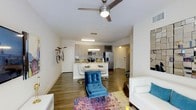 This contemporary single-floor residence showcases a thoughtfully designed open-concept layout that seamlessly connects the living, dining, and kitchen areas into one cohesive, inviting space. Rich hardwood-style flooring runs throughout, anchoring the modern aesthetic and adding warmth to the bright, airy interior.

The living area is a comfortable and stylish retreat, highlighted by a ceiling fan with integrated lighting that provides both function and visual appeal. The space accommodates a relaxed seating arrangement, and a vibrant, multicolored area rug adds a bold pop of color to the neutral palette.

The kitchen is well-appointed with stainless steel appliances, including a French door refrigerator and a microwave, complemented by sleek cabinetry in a warm taupe finish and a tile backsplash. A peninsula counter serves as both a prep space and a casual dining surface, with modern seating positioned along its edge.

Adjacent to the kitchen, the dedicated dining area provides an intimate setting for meals. The clean white walls and neutral tones throughout create a bright and open ambiance, while thoughtful touches — such as wall-mounted décor and vibrant accent furnishings — give the space a personalized, contemporary character. The single-floor plan offers convenient, accessible living with a well-organized flow between all areas.