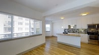 This single-floor space features an open-concept layout that seamlessly connects the living area and kitchen, creating a cohesive and functional environment. Light hardwood flooring runs throughout, lending warmth to the otherwise crisp, contemporary aesthetic defined by clean white walls and white-painted ceilings.

The living area benefits from a large window fitted with horizontal blinds, allowing ample natural light to fill the room while offering views of the surrounding urban landscape, including neighboring buildings and open sky. A second window is visible deeper in the space, further enhancing the bright, airy atmosphere.

The kitchen is well-appointed with white upper cabinetry, a gray mosaic tile backsplash, and a peninsula countertop with a warm-toned surface that provides both workspace and casual seating potential. Pendant lights hang above the peninsula, adding focused task lighting and a modern design touch. The kitchen also includes a built-in stovetop and a recessed ceiling light, completing the functional layout.

Overall, the space presents a clean, modern aesthetic with consistent finishes throughout, making it a well-lit and inviting single-level unit suited for everyday living.