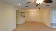 This single-floor residence offers a thoughtfully designed layout centered around a spacious, open-concept living and dining area. The combined living space is finished with plush wall-to-wall carpeting in a warm neutral tone, creating a comfortable and inviting atmosphere throughout.

The dining area is highlighted by an elegant chandelier that serves as a decorative focal point, while a ceiling fan with a built-in light fixture in the adjacent living area ensures year-round comfort and air circulation. Recessed lighting in the ceiling adds a clean, modern touch and provides additional illumination across the space.

A pass-through window connects the kitchen to the main living area, promoting an open feel and easy interaction between spaces. The kitchen, visible through the pass-through and a nearby doorway, features cabinetry and countertops that complement the overall neutral color palette of the home.

The interior is finished with white walls and white baseboards, lending a bright, clean aesthetic that makes the space feel open and airy. The single-floor plan offers a practical and accessible layout, making the home easy to navigate. Overall, the space conveys a welcoming, functional character suited to everyday living and entertaining alike.