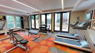 This well-appointed fitness center offers a comprehensive workout environment designed with both functionality and aesthetics in mind. The space is immediately distinguished by its eye-catching geometric-patterned carpet in bold shades of orange, red, and gray, lending the room a dynamic and energetic atmosphere.

The facility is equipped with a well-rounded selection of exercise equipment, including treadmills for cardio training, a multi-functional cable and weight machine along the wall, a dedicated free weights station with a tiered dumbbell rack, and an adjustable workout bench for strength training. The layout ensures ample space between equipment, allowing for comfortable movement throughout the room.

Natural light floods the interior through a series of large, floor-to-ceiling glass-paneled doors with black frames, which open directly onto an outdoor patio. The patio is visible beyond the doors and features seating and what appears to be an outdoor grill, extending the usable amenity space outdoors. Recessed linear LED lighting runs along the ceiling, providing consistent, modern illumination throughout the day and evening. The combination of abundant natural light, a bright color palette, and thoughtful equipment placement creates an inviting and motivating environment for fitness enthusiasts.