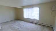 This single-floor space presents a clean and functional layout centered around a spacious main room. The room is finished with wall-to-wall carpeting in a neutral tone, providing a comfortable and quiet underfoot feel throughout. A large picture window serves as the focal point of the room, fitted with vertical blinds that allow for adjustable light control. The window draws in generous amounts of natural light, brightening the interior and creating an open, airy atmosphere. Below the window, a baseboard heater runs along the wall, offering an efficient and unobtrusive heating solution. The walls are finished in a soft, neutral cream tone, complementing the light carpeting and contributing to an overall understated palette. The textured ceiling adds a subtle architectural detail to the otherwise clean interior. The room offers ample open floor space, making it well-suited for a variety of furniture arrangements. The straightforward, no-frills design of the space prioritizes functionality and practicality, providing a versatile canvas for personalization.