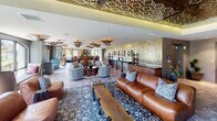 Located at 26600 Oso Pkwy in Mission Viejo, California, this single-floor space is a stunning showcase of Moroccan-inspired interior design. The main lounge area is anchored by a series of ornate, saucer-shaped metallic pendant lights and a dramatic gold mosaic tile accent wall adorned with decorative mirrors and brass fixtures. Rich leather sofas and boldly patterned upholstered swivel chairs are arranged atop layered area rugs, creating an inviting yet sophisticated atmosphere.

A formal dining area features a large, dark wood communal table surrounded by intricately carved, jewel-toned upholstered chairs beneath a striking laser-cut wood ceiling panel with warm ambient uplighting. The hexagonal and geometric patterned tile flooring transitions seamlessly throughout the space, adding visual texture underfoot.

A sunlit circular alcove serves as a standout feature, with a wraparound cushioned banquette, colorful accent pillows, a hand-painted mosaic center table, and a coffered patterned ceiling with a Moroccan lantern pendant. Floor-to-ceiling windows in this nook and throughout the dining area offer panoramic views of lush green landscaping, flooding the interior with natural light. The overall ambiance is warm, eclectic, and richly layered.