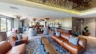 Located at 26600 Oso Pkwy in Mission Viejo, California, this single-floor space is a stunning showcase of Moroccan-inspired interior design. The main lounge area is anchored by a series of ornate, saucer-shaped metallic pendant lights and a dramatic gold mosaic tile accent wall adorned with decorative mirrors and brass fixtures. Rich leather sofas and boldly patterned upholstered swivel chairs are arranged atop layered area rugs, creating an inviting yet sophisticated atmosphere.

A formal dining area features a large, dark wood communal table surrounded by intricately carved, jewel-toned upholstered chairs beneath a striking laser-cut wood ceiling panel with warm ambient uplighting. The hexagonal and geometric patterned tile flooring transitions seamlessly throughout the space, adding visual texture underfoot.

A sunlit circular alcove serves as a standout feature, with a wraparound cushioned banquette, colorful accent pillows, a hand-painted mosaic center table, and a coffered patterned ceiling with a Moroccan lantern pendant. Floor-to-ceiling windows in this nook and throughout the dining area offer panoramic views of lush green landscaping, flooding the interior with natural light. The overall ambiance is warm, eclectic, and richly layered.