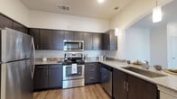 This single-story unit offers a thoughtfully designed layout that maximizes its well-proportioned footprint. The kitchen serves as a standout feature, showcasing dark espresso cabinetry with sleek bar-pull hardware, light granite countertops, and a classic white subway tile backsplash. A full suite of stainless steel appliances — including a refrigerator, range, microwave, and dishwasher — completes the space, while warm wood-style luxury vinyl flooring and a pendant light add a polished, contemporary aesthetic.

The living area transitions seamlessly from the kitchen and features carpet flooring and a ceiling fan. A sliding glass door with vertical blinds opens to a private balcony, providing natural light and outdoor views. The bedroom is carpeted and offers generous wall space, with crown molding adding a refined detail. A door within the bedroom provides convenient access to additional spaces.

The bathroom mirrors the design language of the kitchen, featuring matching dark cabinetry, granite countertops, and wood-style flooring. A shower-tub combination with a curved curtain rod completes the space. Throughout the unit, neutral tones and consistent finishes create a cohesive, modern ambiance.