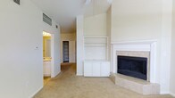 This single-story space makes a strong impression with its dramatically vaulted ceilings, which lend an expansive, open feel to the main living area. The room centers around a tiled fireplace with a classic white mantel surround, providing a focal point that adds warmth and character. Adjacent to the fireplace, a built-in shelving unit with lower cabinet storage offers practical display and organizational space, ideal for media equipment or decorative items.

Soft, neutral-toned carpeting covers the floor throughout, contributing to a comfortable and cohesive aesthetic. The walls are finished in a warm off-white tone, complementing the bright, clean trim work and creating a welcoming atmosphere.

A hallway visible from the main living space leads to additional rooms, including what appears to be a bathroom equipped with vanity lighting and wood-toned cabinetry, as well as a closet space. The layout suggests a functional flow between the living area and private spaces.

Overall, the space is characterized by its generous ceiling height, thoughtful built-in features, and a neutral palette that provides a versatile foundation for a variety of interior styles.