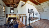 This striking two-floor space occupies a beautifully repurposed historic building that masterfully balances original architectural character with contemporary functionality. The structure's most commanding features are its exposed limestone and plaster walls, warm wood-beam ceilings with visible trusses, and rich terra cotta tile flooring that carry throughout much of the space.

The grand entry welcomes visitors through a stately arched wood door topped with a fanlight window, flooding the foyer with natural light. Original stone walls flank the entryway, setting the tone for the building's storied aesthetic. A wrought iron staircase connects the two levels, offering views across the open floor plan below.

The main floor features dedicated workstations set against textured stone walls, bathed in natural light from large wood-framed windows. Multiple lounge areas are furnished with leather seating, upholstered chairs, and glass coffee tables, creating both collaborative and private retreat spaces. Decorative patterned rugs add warmth and definition to each area.

A coffee bar with a dark wood countertop, reclaimed wood paneling, and pendant lighting provides a communal gathering point. The upper level offers a mezzanine corridor with additional seating and display areas, overlooking the activity below. Throughout, vintage artwork, framed photography, and curated décor reinforce the space's distinctive character.