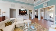 This well-appointed single-floor community clubhouse offers a thoughtfully designed gathering space that balances comfort and functionality. The interior is unified by warm terracotta tile flooring, crisp white trim, and soothing teal accent walls that lend the space a welcoming, resort-inspired ambiance throughout.

The central lounge area serves as the heart of the space, featuring a plush white leather sectional sofa and ottoman arranged atop a colorful area rug. A striking tile-surround fireplace with a white mantel anchors one wall, complemented by a wall-mounted flat-screen television set within a teal-painted niche. A tall floor mirror and decorative accessories add a refined, polished touch.

Adjacent to the lounge, a café-style kitchen bar with dark cabinetry, pendant lighting, and a mosaic tile backsplash provides a convenient refreshment station. The dining area nearby is bathed in abundant natural light streaming through a dramatic wall of oversized windows, offering picturesque views of the outdoor pool and surrounding landscaping. Bistro-height tables paired with cream upholstered chairs create a bright and inviting spot for casual gatherings.

A dedicated business center provides a quiet, carpeted workspace with ample room for productivity. Crown molding throughout and high ceilings enhance the overall sense of openness and elegance within this versatile community space.