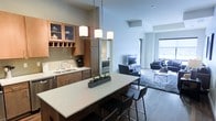 This single-floor space showcases a thoughtfully designed open-concept layout that integrates the kitchen and living area into one cohesive environment. The kitchen is a standout feature, outfitted with warm-toned wood cabinetry, glass-front upper cabinets with built-in wine rack storage, and a subway tile backsplash. A large central island with a white countertop serves as both a prep surface and casual dining space, accommodated by sleek bar-height seating. Pendant lighting with square white shades hangs above the island, providing focused illumination and a modern aesthetic touch. The sink area is equipped with stainless steel fixtures and under-cabinet storage. Seamlessly connecting to the kitchen, the living area features hardwood-style flooring, a plush area rug, and a media setup with comfortable seating arranged around a glass coffee table. Large windows along the far wall allow generous natural light to fill the space, creating a bright and welcoming atmosphere throughout. The open sightlines between zones enhance the sense of spaciousness, making the layout feel expansive and well-suited for both everyday living and entertaining.