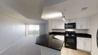 This single-floor space offers a thoughtfully designed open-concept layout that seamlessly connects the kitchen and living area, creating a cohesive and functional living environment. The kitchen is a standout feature, showcasing sleek dark granite countertops that extend across both the prep area and a peninsula breakfast bar. Crisp white cabinetry provides ample storage, while black appliances — including an electric range and over-the-range microwave — add a bold, modern contrast. Track lighting overhead ensures the kitchen workspace is well-lit and practical. The flooring transitions from light hardwood-style material in the kitchen to plush carpeting in the living area, subtly defining each zone while maintaining an open feel. The living area is generously sized and benefits from abundant natural light streaming through a large sliding glass door fitted with vertical blinds, hinting at outdoor access beyond. The textured ceiling and clean white walls throughout lend a neutral, versatile backdrop. Overall, the space presents a balanced blend of contemporary finishes and practical design, making it both visually appealing and highly livable.