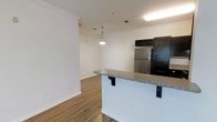 This single-floor space showcases a thoughtfully designed open-concept layout that seamlessly connects the kitchen and living areas. The kitchen is a standout feature, outfitted with rich dark cabinetry, a stainless steel refrigerator, and a peninsula-style breakfast bar topped with granite countertops — ideal for casual dining or entertaining. A stylish pendant light hangs above, adding a warm, focused glow to the space.

The flooring throughout is a warm hardwood-style material that adds continuity and a sense of cohesion across the open floor plan. Crown molding runs along the ceiling, lending an elevated, polished feel to the interior. The walls are finished in clean, bright white, enhancing the sense of openness and allowing natural and artificial light to reflect throughout the space.

The adjacent living area is open and airy, benefiting from the flowing layout and consistent finishes. Visible ceiling vents ensure year-round climate comfort. Overall, the space strikes a balance between functional design and modern aesthetics, with quality finishes and a well-considered layout that maximizes usability and visual appeal.
