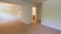 This two-floor space features a straightforward, functional layout with a neutral color palette throughout. The main living area is finished with wall-to-wall beige carpeting, lending a warm and understated feel to the space. Cream-colored walls and standard-height ceilings create a clean, simple backdrop throughout.

An open passageway connects the main living area to the kitchen, which is visible in the background and equipped with white appliances, including a refrigerator, along with warm-toned wooden cabinetry. This open sightline between the living and kitchen areas contributes to an easy flow across the floor plan.

A doorway leads into a bathroom, which features wooden vanity cabinetry and a lighted vanity mirror, providing practical functionality. Adjacent to the bathroom entrance, a closet with a sliding door offers convenient built-in storage.

The space is designed with practicality in mind, offering defined zones for living, cooking, and personal use. The consistent use of neutral tones and carpeted flooring throughout the visible areas creates a cohesive and understated atmosphere across both levels of the home.
