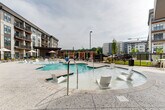 Located at 704 W Tremont Ave in Charlotte's vibrant South End neighborhood, this outdoor amenity space offers a resort-inspired pool experience designed for relaxation and leisure. The centerpiece is a generously sized, freeform swimming pool with a smooth concrete deck surround, complemented by a separate shallow wading pool fitted with modern white in-water lounge chairs — ideal for lounging while staying cool. The pool area is framed by the contemporary mixed-material architecture of the surrounding building, which features a striking combination of brick, board-and-batten siding, and expansive windows with private balconies overlooking the deck below. Sun loungers with navy blue cushions and striped market umbrellas line the perimeter, providing ample space for sunbathing. A pergola structure with string lights anchors a casual seating area, creating an inviting spot for social gatherings. Additional shaded cabanas and umbrella-covered seating areas are distributed throughout the space, offering varied options for relaxation. Lush landscaping, including ornamental grasses and columnar evergreens, adds greenery and privacy. The open-air setting benefits from expansive sightlines and an airy atmosphere, making this a well-appointed outdoor retreat within an urban residential community.