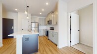 This single-floor space offers a thoughtfully designed layout with a cohesive modern aesthetic throughout. The kitchen serves as a standout feature, showcasing soft gray shaker-style cabinetry with matte black hardware, white quartz countertops, and a full suite of stainless steel appliances, including a refrigerator, electric range, and over-the-range microwave. A central island provides additional prep space and sink access, topped with matching quartz and illuminated by pendant lighting above. White subway tile backsplash and light wood-tone flooring complete the kitchen's clean, contemporary look.

The bedroom is generously proportioned, finished with neutral carpet and painted in warm, soft tones. A large multi-panel window fills the room with natural light and provides exterior views. A walk-in closet with wire shelving systems offers practical storage capacity.

The bathroom features shaker-style cabinetry consistent with the kitchen, a white quartz vanity top, an undermount sink, and matte black fixtures and hardware. Light wood-tone flooring extends through this space, maintaining a unified design language. Recessed lighting and clean white walls contribute to an overall bright, fresh ambiance throughout.
