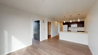 This thoughtfully designed single-floor unit offers a clean, contemporary aesthetic throughout, with light wood-tone flooring and crisp white walls creating a bright, cohesive atmosphere. The open-concept layout centers around a sleek kitchen island featuring a white countertop, undermount sink, and elegant pendant lighting. Dark espresso cabinetry lines the kitchen walls, providing generous storage and a striking contrast against white appliances, including a refrigerator, range, and microwave. The kitchen flows seamlessly into the main living area, making the space ideal for everyday living and entertaining.

Two bedrooms offer peaceful retreats, each featuring large windows that draw in abundant natural light. One bedroom frames sweeping city skyline views, while the other overlooks a lush green treeline, providing a serene urban escape. Both rooms are generously proportioned with ample wall space and hardwood-style flooring throughout.

The bathroom is finished in a modern style, featuring a full soaking tub with white tile surround, a dark vanity with white countertop and single basin sink, and matching brushed nickel hardware. A hallway with a closet connects the living areas to the private rooms, and a frosted glass interior door adds a refined architectural touch to the overall design.