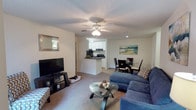 This single-floor residence features two bedrooms and two bathrooms within a thoughtfully designed open-concept floor plan. The living area serves as the central hub of the home, featuring plush carpeted flooring, a ceiling fan with integrated lighting, and a textured ceiling. A large sectional sofa and glass-top coffee table furnish the space comfortably, creating an inviting atmosphere for relaxation.

The living area flows naturally into the dining space, which accommodates a full dining table and chairs, making it well-suited for everyday meals or entertaining. Adjacent to the dining area, the kitchen is defined by a wraparound breakfast bar that provides both a casual eating surface and a visual boundary between spaces. The kitchen is equipped with dark countertops, black appliances including a range and over-the-range microwave, and bright white cabinetry that adds a clean, cohesive look. Recessed lighting illuminates the kitchen area effectively. The open sightlines between all three areas contribute to an airy, connected feel throughout the main living space.