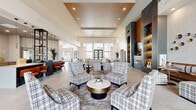 Located at 9600 W Georgia Ave in Glendale, Arizona, this contemporary community clubhouse offers a thoughtfully designed, single-floor space that combines functionality with modern elegance. The main lounge is the centerpiece of the space, boasting soaring double-height ceilings, large floor-to-ceiling windows that flood the interior with natural light, and grand glass-paneled entry doors framing views of the surrounding landscape. The lounge features a striking built-in fireplace flanked by illuminated shelving with dark slate tile surround, a wall-mounted television, and a herringbone-patterned area rug anchoring plush seating arrangements. Decorative metallic wall art and recessed wood ceiling inlays add refined architectural detail throughout. The leasing area includes a sleek white reception counter with pendant lighting, warm amber accent chairs, and an open shelving display partition. A dedicated game room provides entertainment with foosball and billiard tables, a bar-height counter, and textured accent walls beneath a statement chandelier. The catering kitchen is outfitted with wraparound dark countertops, light wood cabinetry, a subway tile backsplash, built-in appliances, and dramatic textured wall treatments. Large-format light tile flooring unifies the entire space, creating a cohesive and polished aesthetic throughout.