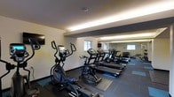 This single-floor fitness space is designed to support a well-rounded cardio workout in a clean, functional environment. The room is furnished with multiple treadmills, an elliptical trainer, and a stationary bike, all arranged to maximize floor space and ease of movement between equipment. Dark rubber flooring mats are positioned beneath the machines, providing stability and floor protection during use. A large wall mirror spans one side of the room, visually expanding the space and allowing users to monitor their form while exercising. Recessed linear lighting runs along the ceiling perimeter, casting an even, warm glow throughout the room. A wall-mounted flat-screen television is positioned at the front of the space, offering entertainment during workouts. A second television is visible reflected in the mirror, ensuring good visibility from multiple vantage points. Natural light filters in through windows along the wall, contributing to a brighter, more open atmosphere. A dumbbell rack is visible near the mirrored wall, adding a modest free-weight option to complement the cardio equipment. The textured ceiling and neutral wall tones give the space a practical, no-frills aesthetic focused on fitness functionality.