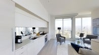 This single-floor space showcases a contemporary open-plan design that seamlessly integrates the kitchen and living areas. The kitchen is outfitted with handleless white cabinetry that runs the full length of the wall, complemented by a mirrored backsplash that amplifies natural light and adds visual depth. Stainless steel appliances, including a gas cooktop and built-in oven, are integrated cleanly into the cabinetry, maintaining the minimalist aesthetic. A undermount sink with modern fixtures completes the functional workspace.

The living area benefits from floor-to-ceiling windows and a sliding glass door that floods the interior with abundant natural light while framing sweeping elevated views of the city skyline and tree canopy beyond. Light-toned hardwood flooring runs throughout, reinforcing the bright, airy atmosphere. The neutral white palette across walls and ceilings enhances the sense of openness and scale. A sliding glass door provides access to an outdoor balcony, further connecting the interior to the panoramic surroundings. The overall ambiance is clean, modern, and light-filled.