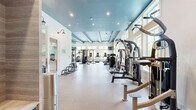 Located at 2200 Flagler Promenade Way in Maitland, Florida, this fitness center is a generously sized, single-floor facility divided into two dedicated exercise rooms, offering a comprehensive workout experience. The main exercise room features soaring ceilings with bold dark-painted exposed beams, recessed lighting, and large ceiling fans, creating an energizing yet comfortable atmosphere. An extensive array of strength training equipment fills the space, including a cable machine, power rack with free weights, a heavy bag for boxing, and multiple resistance machines. Treadmills, rowing machines, and other cardio equipment are also available. Full-length mirrors amplify the sense of space, while large windows along the perimeter flood the room with natural light and offer views of the surrounding Florida greenery. The second exercise room provides a more intimate setting with wood-look flooring, a spin bike, a flat bench, and a wall-mounted television — ideal for focused cardio or stretching sessions. A dedicated free weights area features a full dumbbell rack alongside medicine balls and stability equipment. Throughout both rooms, floor-to-ceiling windows frame views of palm trees and the outdoor pool area, seamlessly connecting the indoor fitness environment with the natural surroundings outside.