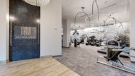 Located at 4401 Barclay Downs Dr in Charlotte, NC, this Wellness Center is a thoughtfully designed, single-floor facility offering a comprehensive health and fitness experience. The entrance welcomes visitors with soaring ceilings, light wood-look flooring, and floor-to-ceiling glass doors that flood the space with natural light and provide views of lush outdoor greenery. A striking reception area features a bold dark stone accent wall bearing the Wellness Center insignia, flanked by decorative backlit cylindrical columns and an intricate laser-cut metal screen partition atop a stone-faced reception desk, establishing an upscale first impression. The expansive main fitness floor is anchored by dramatic oversized circular pendant lights and showcases a full array of cardio equipment, including treadmills and strength machines, set against a sweeping artistic mural wall. Large black-framed windows further connect the space to the outdoors. A dedicated studio room, enclosed by steel-framed glass partitions and accented with exposed wood ceiling beams, accommodates cycling bikes and a weight bench alongside a wall-mounted display. A private, serene massage room with neutral tones and curated wall art rounds out the facility, offering a tranquil retreat for recovery and relaxation.