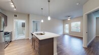 This well-appointed single-floor residence offers a thoughtfully designed open-concept layout that seamlessly connects the kitchen, dining, and living areas. The kitchen serves as a central highlight, anchored by a generously sized curved island with a quartz countertop, undermount sink, and stylish pendant lighting overhead. Rich, dark cabinetry provides ample storage, while a full suite of stainless steel appliances—including a gas range, microwave, and refrigerator—adds a polished, modern touch. A subway tile backsplash and track lighting complete the culinary space. The open living area flows naturally from the kitchen, featuring warm wood-look flooring, neutral toned walls, and a ceiling fan that enhances comfort. A door leads to the outdoors, and multiple windows fitted with horizontal blinds allow for generous natural light throughout. The bedrooms are spacious and serene, each with the same warm flooring and large windows that brighten the rooms. The bathroom is finished with elegant marble-style tile flooring, a glass-enclosed shower, a wall-mounted cabinet with dark wood finish, and clean, neutral walls. Closet space is visible from several rooms, adding to the unit's practical storage capacity. The overall ambiance is clean, contemporary, and move-in ready.