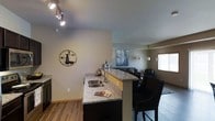 This single-floor space showcases an open-concept design that integrates the kitchen, dining, and living areas into one cohesive environment. The kitchen is anchored by a substantial peninsula topped with speckled granite countertops, offering both prep space and seating along one side with stylish black upholstered bar stools featuring nailhead trim detail. A stainless steel undermount sink is built into the peninsula, complemented by dark cabinetry below. Track lighting on the ceiling provides focused illumination throughout the kitchen area. The flooring is light-toned hardwood, adding warmth and continuity across the open space. Neutral gray walls create a clean, modern backdrop throughout. Transitioning into the living and dining area, the space maintains an airy, open feel with additional seating visible and wall art providing decorative character. A window in the background allows natural light to filter in. The overall ambiance is contemporary and functional, with thoughtful design choices that balance practicality with a polished, inviting aesthetic suited for both everyday living and entertaining.