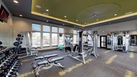 This contemporary fitness center is thoughtfully designed to support a comprehensive workout experience across a single, open floor. The space is anchored by a striking tray ceiling finished in a bold olive-green tone, accented by warm LED lighting that creates a vibrant yet focused atmosphere. Recessed lighting and a ceiling fan complement the elevated ceiling design, contributing to an airy and comfortable environment throughout.

Large floor-to-ceiling windows line multiple walls, flooding the space with natural light and offering outdoor views that enhance the sense of openness. The flooring consists of patterned carpet tiles in charcoal and gold tones, adding a modern, textural element to the overall aesthetic.

The fitness floor is divided into functional zones. The strength training area features a multi-station cable machine, adjustable weight benches, a full rack of dumbbells, and stability balls, all positioned in front of full-length mirrors that visually expand the space and allow for form monitoring. The cardio section is equipped with multiple treadmills, a stair climber, stationary bikes, and a rowing machine, all facing expansive windows. Wall-mounted televisions are positioned throughout for entertainment. Vibrant nature-inspired artwork adds a finishing touch to the overall design.