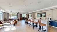 Located at 1100 Cricket Ln in East Walpole, Massachusetts, this single-floor amenity space offers a generously sized, thoughtfully designed environment ideal for socializing and relaxation. The living room is an immediate showpiece, featuring dramatic navy blue paneled walls, a central fireplace with a textured tile surround, a wall-mounted TV, and twin geometric gold-framed pendant lights that lend a refined, contemporary elegance. Plush sofas and sleek marble-topped coffee tables with brass accents complete the sophisticated seating arrangement. Adjacent to the living room, the dining area is bathed in natural light streaming through expansive floor-to-ceiling windows that frame lush outdoor views. A warm wood dining table paired with cognac leather chairs sits beneath a matching gold-framed pendant light, while herringbone-patterned flooring flows throughout, unifying the space. Double glass doors provide direct access to an outdoor terrace. A dedicated billiards area features a full-size pool table set against more large windows overlooking greenery, complemented by leather lounge chairs and a relaxed seating arrangement. A full bar with counter seating and cabinetry in coordinating navy tones anchors the space, reinforcing the cohesive, upscale aesthetic found throughout.