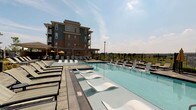 This amenity space is designed to offer both indoor comfort and outdoor recreation. The centerpiece of the outdoor area is a generously sized swimming pool, equipped with sleek in-water lounge chairs that sit partially submerged for a resort-style experience. Surrounding the pool is a spacious deck lined with traditional chaise lounges, a shaded pergola structure with striped canopy accents, and a wrought-iron perimeter fence that frames open views of the surrounding landscape. A multi-story residential building serves as a prominent backdrop, adding a sense of scale to the outdoor setting.

Inside, the clubroom exudes a refined yet welcoming atmosphere. The space is anchored by a floor-to-ceiling stone fireplace with a rustic wood mantel, providing a striking focal point. Neutral-toned sofas and wingback chairs are arranged to encourage conversation and relaxation. Elegant multi-bulb chandeliers illuminate the high ceilings, while large French doors invite natural light and connect the interior to the outdoor pool area. Additional seating nooks and connecting rooms extend the functionality of this well-designed communal space.