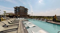 This amenity space is designed to offer both indoor comfort and outdoor recreation. The centerpiece of the outdoor area is a generously sized swimming pool, equipped with sleek in-water lounge chairs that sit partially submerged for a resort-style experience. Surrounding the pool is a spacious deck lined with traditional chaise lounges, a shaded pergola structure with striped canopy accents, and a wrought-iron perimeter fence that frames open views of the surrounding landscape. A multi-story residential building serves as a prominent backdrop, adding a sense of scale to the outdoor setting.

Inside, the clubroom exudes a refined yet welcoming atmosphere. The space is anchored by a floor-to-ceiling stone fireplace with a rustic wood mantel, providing a striking focal point. Neutral-toned sofas and wingback chairs are arranged to encourage conversation and relaxation. Elegant multi-bulb chandeliers illuminate the high ceilings, while large French doors invite natural light and connect the interior to the outdoor pool area. Additional seating nooks and connecting rooms extend the functionality of this well-designed communal space.
