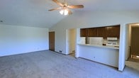 This single-floor space offers a practical and open layout designed for comfortable everyday living. The main living area is a standout feature, boasting vaulted ceilings that lend a sense of spaciousness and airiness to the room. A ceiling fan is centrally mounted, providing both function and a focal point overhead. Soft, neutral-toned carpeting covers the living area floor, contributing to a warm and inviting ambiance.

Adjacent to the living space is a kitchen area, partially open to the main room via a raised breakfast bar or pass-through counter. The kitchen is equipped with dark wood cabinetry that contrasts with the light-colored walls and white appliances, including a refrigerator and range, creating a classic and functional cooking space.

The floor plan also includes a staircase, visible in the overhead view, suggesting access to additional levels or a basement. Multiple doorways lead to additional rooms, including what appears to be a closet or utility space. The overall interior features white walls throughout, offering a clean, neutral backdrop. The layout flows logically between spaces, making the home feel well-organized and easy to navigate.