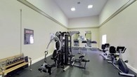 This fitness center offers a comprehensive workout environment designed to accommodate a variety of exercise needs. The space features soaring ceilings that create an open, airy atmosphere, complemented by recessed and panel lighting that ensures the room is well-lit throughout. The walls are finished in a clean, neutral tone and adorned with large mirrors that visually expand the space while allowing users to monitor their form during workouts. The flooring consists of durable rubber matting, providing both safety and comfort underfoot. At the heart of the room stands a robust multi-station strength training machine, offering a wide range of resistance-based exercises suitable for full-body conditioning. Alongside it, a seated cable machine and additional strength equipment provide further options for targeted muscle training. The cardio section is equipped with treadmills and a rowing machine, catering to endurance and low-impact training alike. Motivational fitness posters are displayed along the walls, adding an energetic touch to the environment. The overall layout is thoughtfully organized to allow ample space for movement between equipment, making this fitness center a functional and inviting space for all levels of fitness enthusiasts.