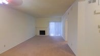 This single-story space presents a straightforward and functional layout centered around a generously sized living area. The room is finished with wall-to-wall carpeting in a neutral beige tone, contributing to a warm and understated atmosphere throughout. A built-in fireplace serves as a focal point along one wall, framed by a simple white mantel that adds a modest architectural detail to the otherwise clean interior. Adjacent to the fireplace, a large window fitted with vertical blinds allows natural light to filter into the room, brightening the space while maintaining privacy. The walls are painted in a soft, neutral hue and are complemented by a textured ceiling, consistent with classic residential construction. Crown molding accents the upper perimeter of the living area, adding a subtle decorative touch. A doorway visible along one wall suggests connectivity to additional areas of the home. The overall ambiance is calm and move-in ready, offering a blank canvas well-suited for a variety of furnishing styles and personal touches.