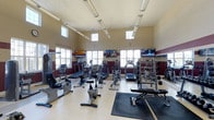Located at 11289 E Hamilton Crest Blvd in Fishers, Indiana, this well-appointed fitness center offers a comprehensive workout environment designed to accommodate a wide range of exercise needs. The space is housed within a single-floor facility and is distinguished by its soaring ceilings and abundant natural light, which streams in through multiple large windows positioned at both standard and clerestory heights, creating an open and energizing atmosphere.

The interior features a clean, neutral color palette accented by deep maroon wall banding, lending the space a structured and purposeful aesthetic. Overhead, linear fluorescent lighting supplements the natural light to ensure consistent visibility throughout all hours of operation.

The facility is generously equipped with cardio machines including treadmills, upright and recumbent stationary bikes, ellipticals, and stair climbers. A dedicated strength training area features a dumbbell rack with a full weight selection and an adjustable workout bench, catering to both beginners and experienced lifters. Multiple wall-mounted flat-screen televisions are positioned throughout the room, allowing members to stay entertained during their workouts. Motivational text displayed on the walls further contributes to an encouraging and focused fitness environment. Rubber floor mats in the free weights area provide safety and durability.