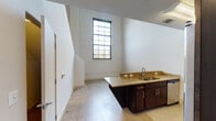 This two-floor loft-style space showcases a compelling blend of industrial heritage and contemporary finishes. The main level features a kitchen equipped with rich dark-stained cabinetry, granite countertops, stainless steel appliances including an electric range and over-the-range microwave, and a double-basin sink set into a peninsula. Polished concrete-style flooring runs throughout the lower level, complementing the exposed painted brick walls that lend unmistakable character to the space. A dramatic tall window on the main floor floods the area with natural light, framing views of the surrounding neighborhood. The bathroom is fitted with a granite-topped vanity, matching dark cabinetry, and a full tub-shower combination, maintaining a cohesive design palette. Upstairs, the upper floor continues the loft aesthetic with soaring ceilings, a whitewashed exposed brick accent wall, and a modern ceiling fan. Gridded black-framed windows allow ample daylight to pour in. The carpeted upper-level bedrooms offer a softer contrast to the industrial elements below, each featuring walk-in closets with built-in shelving. The overall ambiance is open, airy, and distinctly urban, with raw architectural details harmonizing seamlessly with polished, move-in-ready finishes throughout.