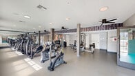 Located at 384 Amwell Rd in Hillsborough, NJ, this single-floor fitness center offers a generously sized, open exercise space designed to support a well-rounded workout experience. The layout is thoughtfully organized, providing dedicated zones for cardio, strength training, and functional fitness without feeling crowded.

The cardio area is well-equipped with multiple SportsArt treadmills, upright and recumbent bikes, ellipticals, and a stair climber, all positioned to take advantage of natural light streaming through a row of large windows along one wall. Wall-mounted flat-screen televisions are conveniently placed for use during cardio sessions. Ceiling fans throughout the space contribute to a comfortable workout environment.

The strength training section features a cable machine, adjustable benches, a dumbbell rack, and stability balls neatly stored in open shelving units. Structural columns clad in a stone-like finish add an architectural element while defining different zones within the open floor plan.

A standout design feature is the bold, large-scale typographic mural covering one full wall, displaying fitness-themed words such as "Workout," "Nutrition," "Fitness," and "Athlete," creating a motivating and energetic ambiance. The light gray speckled flooring, white ceilings with recessed lighting, and large mirrors further enhance the bright, clean aesthetic of the space. A separate room with glass-panel sliding doors provides additional functional space within the facility.