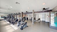 Located at 384 Amwell Rd in Hillsborough, NJ, this single-floor fitness center offers a generously sized, open exercise space designed to support a well-rounded workout experience. The layout is thoughtfully organized, providing dedicated zones for cardio, strength training, and functional fitness without feeling crowded.

The cardio area is well-equipped with multiple SportsArt treadmills, upright and recumbent bikes, ellipticals, and a stair climber, all positioned to take advantage of natural light streaming through a row of large windows along one wall. Wall-mounted flat-screen televisions are conveniently placed for use during cardio sessions. Ceiling fans throughout the space contribute to a comfortable workout environment.

The strength training section features a cable machine, adjustable benches, a dumbbell rack, and stability balls neatly stored in open shelving units. Structural columns clad in a stone-like finish add an architectural element while defining different zones within the open floor plan.

A standout design feature is the bold, large-scale typographic mural covering one full wall, displaying fitness-themed words such as "Workout," "Nutrition," "Fitness," and "Athlete," creating a motivating and energetic ambiance. The light gray speckled flooring, white ceilings with recessed lighting, and large mirrors further enhance the bright, clean aesthetic of the space. A separate room with glass-panel sliding doors provides additional functional space within the facility.