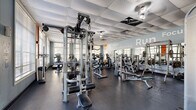Located at 8151 Patterson Woods Dr in Orlando, FL, this fitness center offers a thoughtfully organized exercise space across a single floor. The room is finished with a drop-panel ceiling, resilient rubber flooring, and soft blue-gray walls adorned with large motivational typography — including words like "Run," "Lift," "Power," "Commitment," "Focus," "Energy," "Goals," and "Fitness" — creating an energizing atmosphere throughout.

Natural light streams in through multiple large windows fitted with horizontal blinds, providing views of the surrounding greenery and keeping the space feeling open and airy. Full-length mirrors along the walls further enhance the sense of depth and allow users to monitor their form during workouts.

The cardio section is well-stocked with Life Fitness treadmills, elliptical trainers, upright and recumbent stationary bikes, and stair climbers. The strength training area features a multi-station cable machine, a variety of weight benches, squat racks, and a full dumbbell rack with a wide range of free weights. A wall-mounted flat-screen television adds convenience for entertainment during workouts. The space is designed to accommodate a broad range of fitness routines, from high-intensity cardio sessions to comprehensive strength training programs.