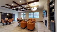 This single-floor commercial space combines contemporary design with warm, inviting aesthetics to create a professional and comfortable environment. The main lounge area is anchored by a striking coffered ceiling with dark wood beam accents, complemented by a large pendant light fixture and recessed lighting. Light hardwood flooring runs throughout, adding warmth and continuity to the space. Comfortable seating arrangements feature rich amber-toned armchairs and patterned sofas, creating an inviting gathering area. Large windows along the perimeter allow natural light to flow in and offer views of the surrounding exterior. A wall-mounted flat-screen display and a sliding barn door add functional and stylistic detail to the lounge.

A glass-walled conference room provides a visually open yet acoustically private meeting space. Inside, a round table is surrounded by rolling amber chairs, encouraging collaborative discussion. The room is distinguished by a wood-paneled ceiling, a stacked stone accent wall with a wall-mounted display, and sophisticated track and recessed lighting. Adjacent to the conference room, a luminous reception or service counter is visible, reinforcing the professional character of the space. Together, these areas reflect a cohesive design philosophy that balances functionality with refined, contemporary style.
